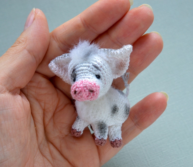 May include: A small crocheted pig toy with white and gray fur, pink ears, and a pink snout. The pig is sitting in a hand.