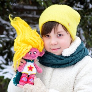 Peut inclure: Une jeune personne portant un bonnet jaune en tricot et une écharpe verte tient une poupée troll en crochet rose aux cheveux jaunes. La poupée porte une chemise blanche avec une étoile et des bottes roses.