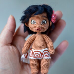 K&ouml;nnte beinhalten: Eine geh&auml;kelte Puppe mit brauner Haut, schwarzen Haaren und gro&szlig;en braunen Augen. Die Puppe tr&auml;gt einen wei&szlig;en Rock mit einem rot-wei&szlig;en Muster. Die Puppe h&auml;lt eine rosa Blume in ihren Haaren.