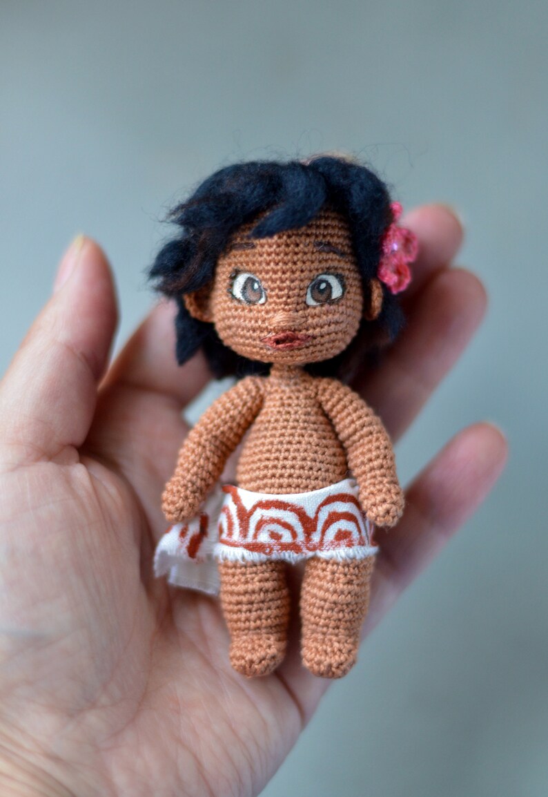 K&ouml;nnte beinhalten: Eine geh&auml;kelte Puppe mit brauner Haut, schwarzen Haaren und gro&szlig;en braunen Augen. Die Puppe tr&auml;gt einen wei&szlig;en Rock mit einem rot-wei&szlig;en Muster. Die Puppe h&auml;lt eine rosa Blume in ihren Haaren.