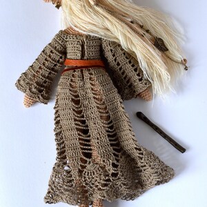 K&ouml;nnte beinhalten: Handgefertigte H&auml;kelpuppe mit langem, flie&szlig;endem blondem Haar und einem braunen Kleid. Die Puppe hat einen braunen G&uuml;rtel und eine kleine Holzfl&ouml;te. Das Kleid der Puppe hat ein einzigartiges Lochmuster.
