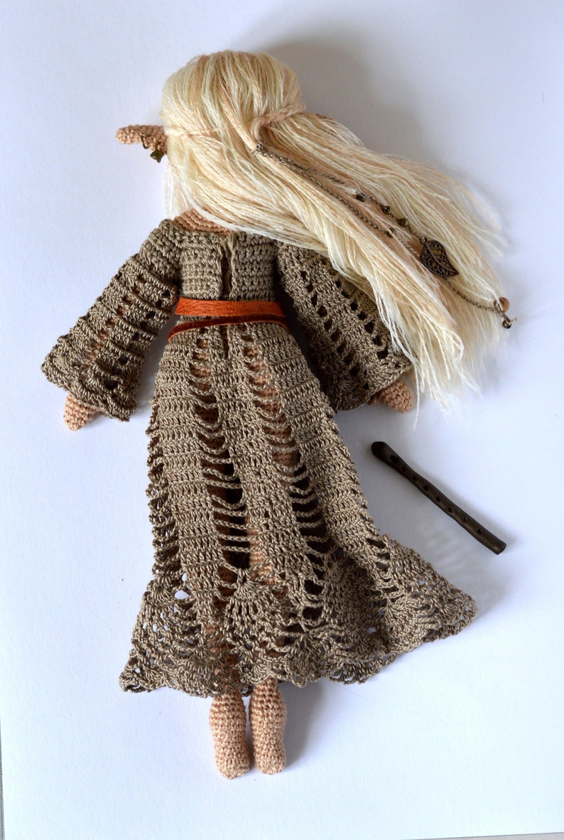 K&ouml;nnte beinhalten: Handgefertigte H&auml;kelpuppe mit langem, flie&szlig;endem blondem Haar und einem braunen Kleid. Die Puppe hat einen braunen G&uuml;rtel und eine kleine Holzfl&ouml;te. Das Kleid der Puppe hat ein einzigartiges Lochmuster.