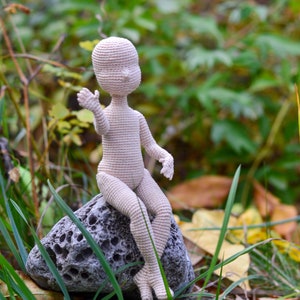 Può includere: Una bambola all'uncinetto con un corpo rosa chiaro seduta su una roccia grigia con erba verde e foglie marroni sullo sfondo.
