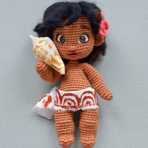 K&ouml;nnte beinhalten: H&auml;kelpuppe mit brauner Haut, schwarzen Haaren und einer Muschel in der Hand. Sie tr&auml;gt einen wei&szlig;en Rock mit roten und orangen Mustern.