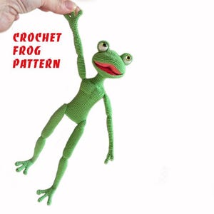 May include: A green crocheted frog with large, round eyes and a red mouth. The frog is suspended in the air, with one arm raised. The text "CROCHET FROG PATTERN" is in red letters on the left side of the image.