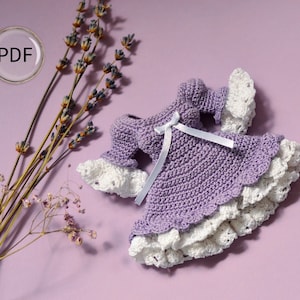 Op de afbeelding: Een paarse gehaakte jurk met witte kant en een wit lint. De jurk ligt op een lichtpaarse achtergrond met gedroogde lavendelbloemen.