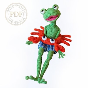 May include: A green crocheted frog doll wearing blue and white striped shorts and an orange crab-shaped swim ring. The frog has large eyes and a pink mouth. A circular badge in the top left corner contains the text Digital Crochet Pattern PDF.