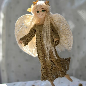 K&ouml;nnte beinhalten: Eine handgefertigte H&auml;kelpuppe in Form eines Engels mit langem, flie&szlig;endem blondem Haar und gr&uuml;nen Augen. Die Puppe tr&auml;gt ein goldfarbenes Kleid und hat gro&szlig;e, spitzenartige Fl&uuml;gel. Kleine, goldfarbene Schmetterlingsakzente schm&uuml;cken den Kopf der Puppe. Die Puppe wird auf einem transparenten St&auml;nder ausgestellt.