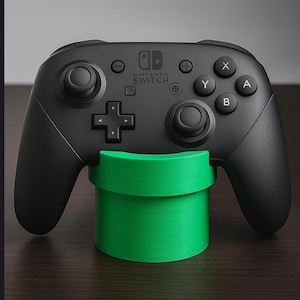 Puede incluir: Un mando negro Nintendo Switch Pro apoyado en un soporte cilíndrico verde brillante. El mando tiene el logotipo de Nintendo Switch y los botones estándar. El soporte se asemeja a una tubería verde.