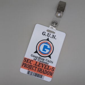 Könnte beinhalten: Weißer Plastik-Ausweis mit einem blauen und orangefarbenen Logo, auf dem "G.U.N. Guardian Units of Nations" und "SEC. LEVEL-5 PROJECT SHADOW" steht.