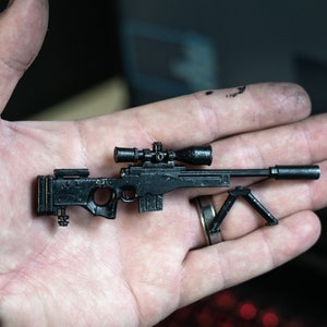 Peut inclure: Un fusil de sniper miniature noir avec une lunette et un bipied. Le fusil est tenu dans une main, mettant en valeur sa petite taille et ses détails complexes. La lunette est noire et cylindrique, et le bipied est un petit support triangulaire.