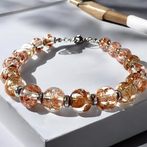 Puede incluir: Un brazalete con cuentas redondas y transparentes con inclusiones de color cobre. Las cuentas están ensartadas en una cadena plateada y aseguradas con un cierre. El brazalete se exhibe sobre una superficie blanca.