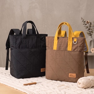 Puede incluir: Dos mochilas rectangulares, una negra y otra marrón, con bolsillos delanteros acolchados, asas superiores dobles y correas ajustables. La mochila marrón tiene asas de color amarillo brillante y una etiqueta rectangular con el nombre Emma.