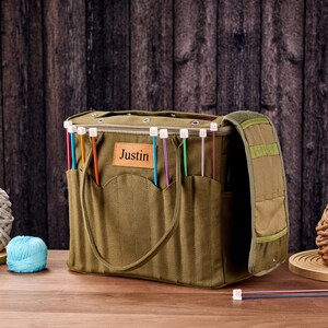 May include: An olive green knitting bag with pockets, a shoulder strap, and the name "Justin" on a leather patch. The bag contains colourful knitting needles. Several balls of yarn in various colours are also present.