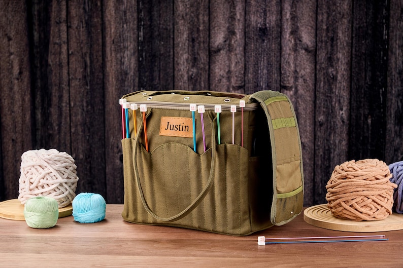 May include: An olive green knitting bag with pockets, a shoulder strap, and the name "Justin" on a leather patch. The bag contains colourful knitting needles. Several balls of yarn in various colours are also present.