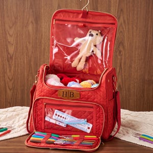 Peut inclure: Un sac de rangement matelassé rouge doté d'un crochet de suspension, d'une poche intérieure en vinyle transparent contenant un ours en peluche et d'une poche avant avec fenêtre transparente affichant une règle. Il inclut des crochets colorés.