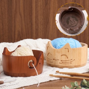 May include: Two wooden yarn bowls, one dark brown and one light tan, featuring a cat face design with a spiral cutout for yarn. The dark bowl contains a white yarn ball, while the light bowl holds a blue yarn ball. Two wooden crochet hooks are visible.