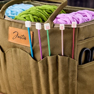 May include: An olive green canvas yarn tote bag with multiple pockets, holding blue, green, and purple yarn. The bag has a leather patch with the name "Justin" and various knitting needles, including 2.0 mm, 3.5 mm, and 4.0 mm sizes.