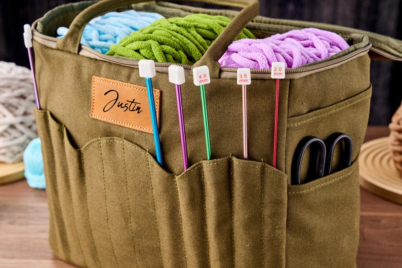 May include: An olive green canvas yarn tote bag with multiple pockets, holding blue, green, and purple yarn. The bag has a leather patch with the name "Justin" and various knitting needles, including 2.0 mm, 3.5 mm, and 4.0 mm sizes.