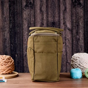 May include: An olive green craft bag with multiple pockets and a zip closure. The bag is surrounded by balls of yarn in various colours, including beige, white, blue, and green. Knitting needles are visible on the left.
