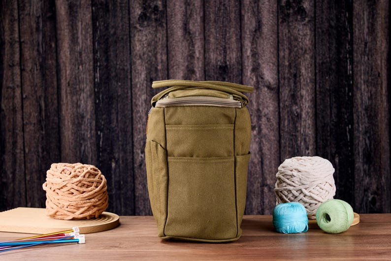 May include: An olive green craft bag with multiple pockets and a zip closure. The bag is surrounded by balls of yarn in various colours, including beige, white, blue, and green. Knitting needles are visible on the left.
