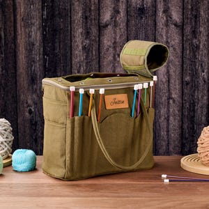 May include: An olive green knitting bag with multiple pockets and a handle. The bag is filled with colorful knitting needles. A small leather patch with the name "Justin" is on the front. Yarn balls and knitting needles are on the wooden surface.