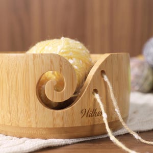 May include: A light-colored wooden yarn bowl with a smooth finish and a spiral cutout for feeding yarn. The bowl features two small circular holes on the side and the word "Wither" engraved in a cursive script. A ball of yellow yarn sits inside.