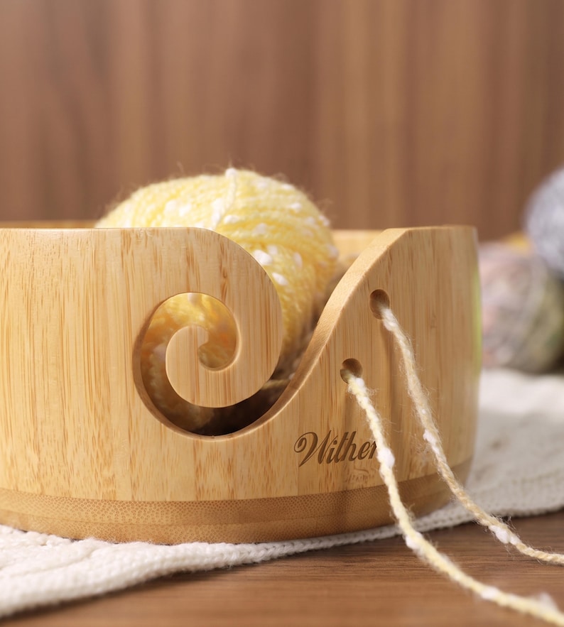 May include: A light-colored wooden yarn bowl with a smooth finish and a spiral cutout for feeding yarn. The bowl features two small circular holes on the side and the word "Wither" engraved in a cursive script. A ball of yellow yarn sits inside.