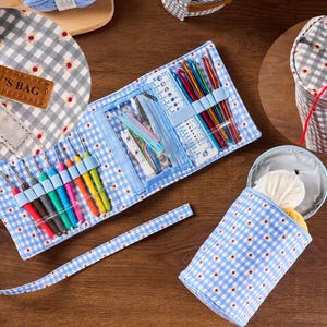 May include: A blue and white gingham crochet hook case and yarn holder set. The case is open, revealing colorful crochet hooks and tools. The yarn holder is cylindrical. The set includes a label that says "NANCY'S BAG".