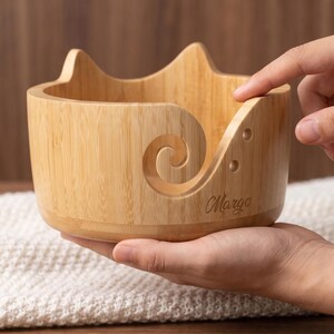 May include: A light-colored wooden yarn bowl featuring a cat-ear silhouette on the top rim and a spiral cutout on the side for feeding yarn. The bowl has two small circular holes near the spiral and the name Margo engraved on the front surface.