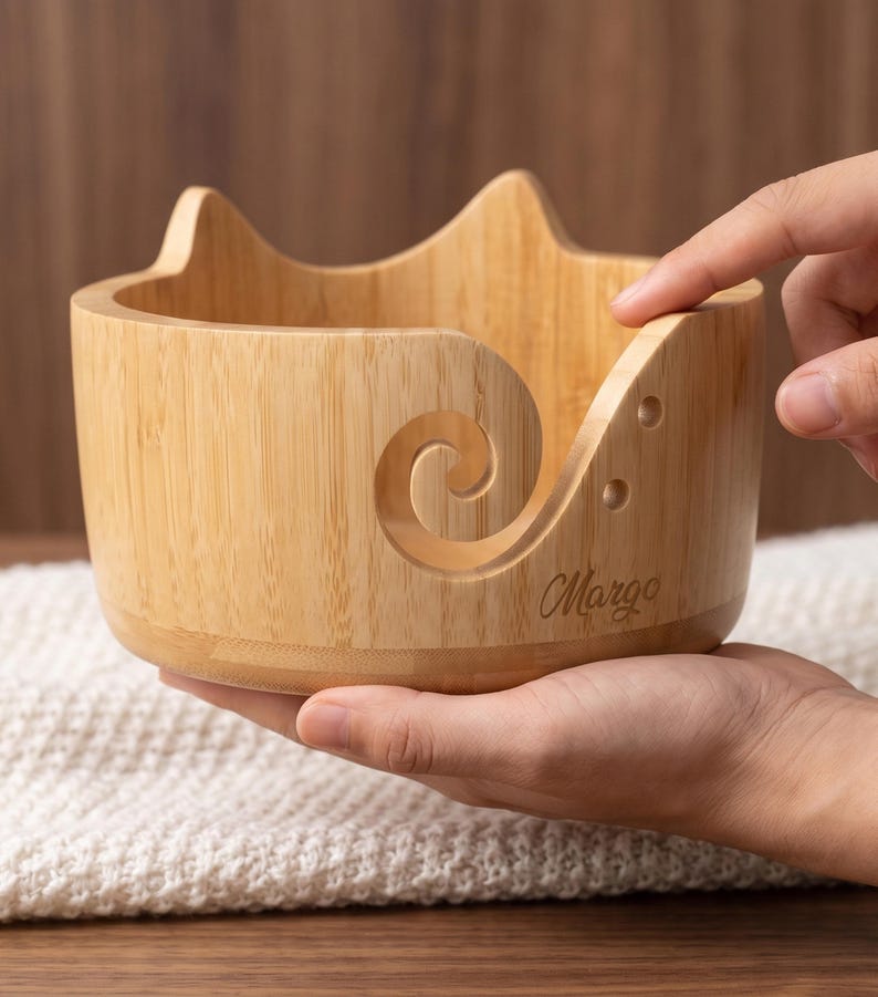 May include: A light-colored wooden yarn bowl featuring a cat-ear silhouette on the top rim and a spiral cutout on the side for feeding yarn. The bowl has two small circular holes near the spiral and the name Margo engraved on the front surface.
