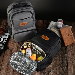 May include: A grey backpack and a black backpack with an open insulated compartment containing glass bottles, a Budweiser can, and fresh fruit. Both backpacks feature a brown leather-like patch on the front exterior pocket.