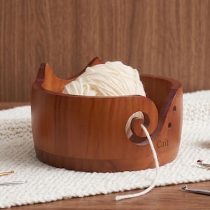 May include: A round, medium-brown wooden yarn bowl featuring two pointed cat-like ears on the rim and a spiral cutout for feeding yarn. The name Cait is engraved on the side. A ball of cream-colored yarn sits inside, with a strand threaded through the cutout.