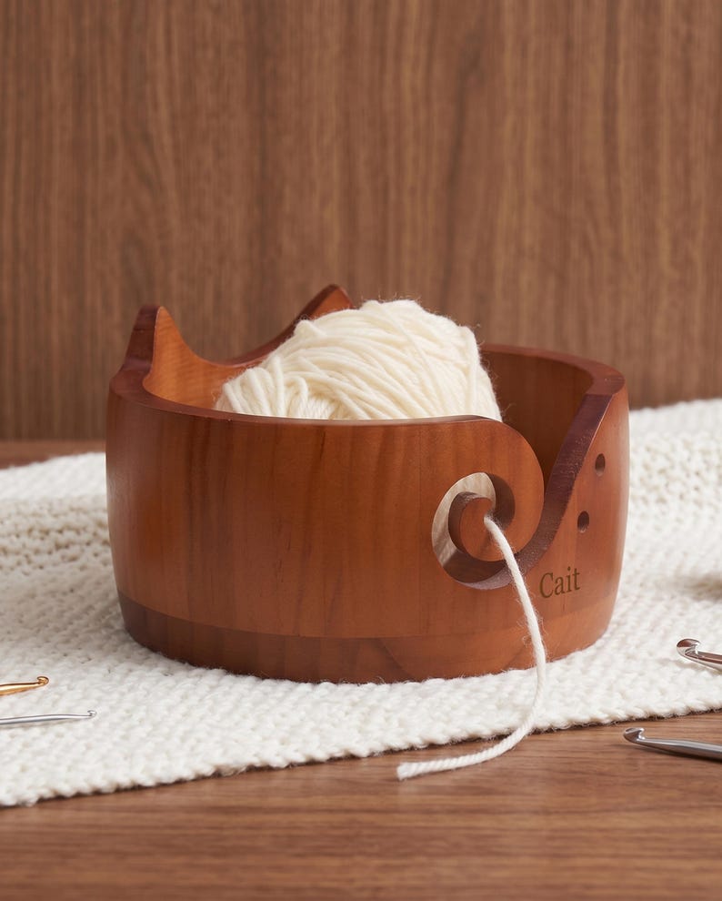 May include: A round, medium-brown wooden yarn bowl featuring two pointed cat-like ears on the rim and a spiral cutout for feeding yarn. The name Cait is engraved on the side. A ball of cream-colored yarn sits inside, with a strand threaded through the cutout.
