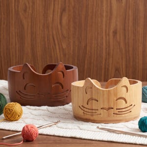 May include: Two round wooden yarn bowls featuring a carved cat face design with pointed ears, closed eyes, and whiskers. One bowl is a dark brown wood, and the other is a lighter natural bamboo color. Both bowls have a curved opening for yarn.
