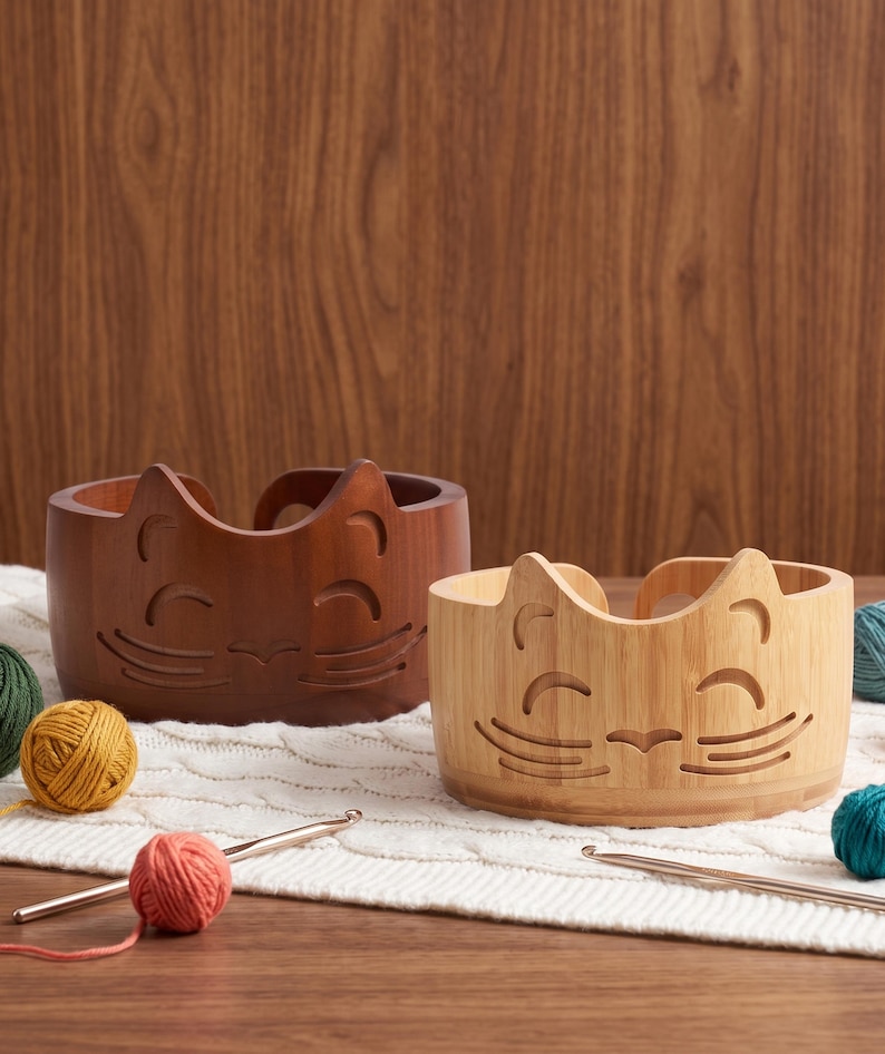 May include: Two round wooden yarn bowls featuring a carved cat face design with pointed ears, closed eyes, and whiskers. One bowl is a dark brown wood, and the other is a lighter natural bamboo color. Both bowls have a curved opening for yarn.