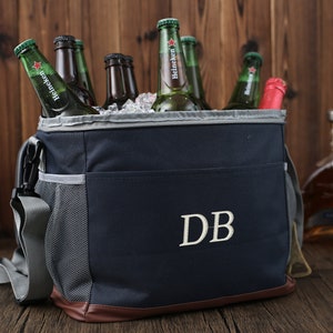 May include: A navy blue insulated cooler bag with a brown leather trim and a gray mesh pocket. The bag has a zipper closure and a shoulder strap. The bag is filled with ice and bottles of beer. The bag has the initials "DB" embroidered on the front.