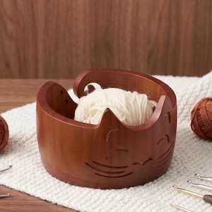 May include: A round, dark brown wooden yarn bowl featuring a carved cat face design with pointed ears and whiskers. The bowl has a spiral cutout on the side for feeding yarn and contains a ball of cream-colored yarn inside the hollow center.