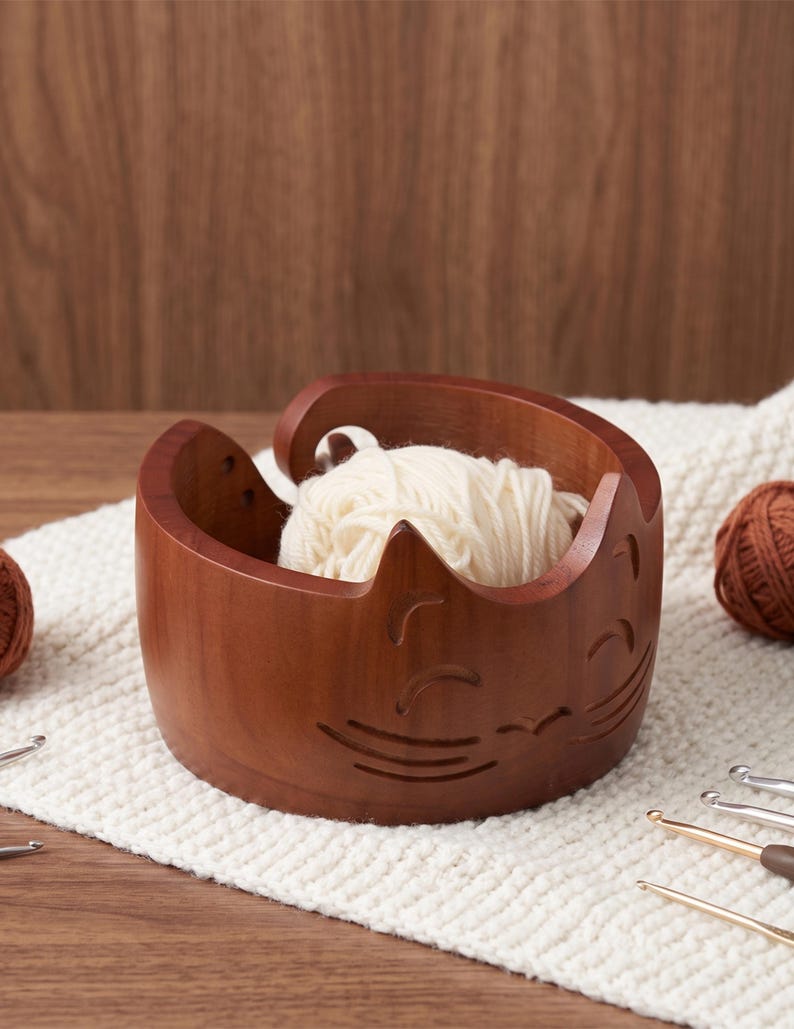 May include: A round, dark brown wooden yarn bowl featuring a carved cat face design with pointed ears and whiskers. The bowl has a spiral cutout on the side for feeding yarn and contains a ball of cream-colored yarn inside the hollow center.