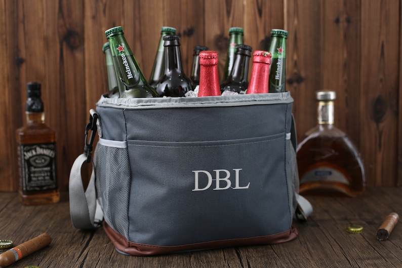 May include: A gray insulated cooler bag with a brown leather trim and a zippered pocket. The bag has a shoulder strap and the initials "DBL" embroidered on the front. The bag is filled with various bottles of beer and wine.