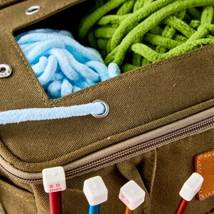 May include: An olive green canvas knitting bag with a zip and a leather patch. The bag contains blue, green, and purple yarn. Knitting needles with white tops and red and blue shafts are visible.
