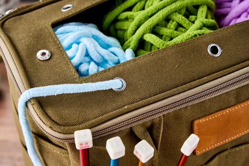 May include: An olive green canvas knitting bag with a zip and a leather patch. The bag contains blue, green, and purple yarn. Knitting needles with white tops and red and blue shafts are visible.