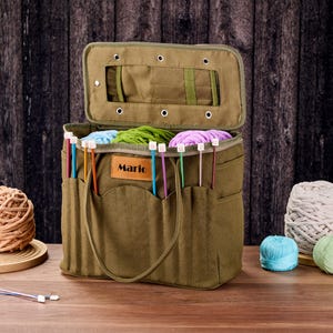May include: An olive green canvas knitting bag with an open lid, filled with colourful yarn and knitting needles. The bag has exterior pockets and a handle. The word "Mario" is on a leather tag on the front. Yarn balls are on the table.
