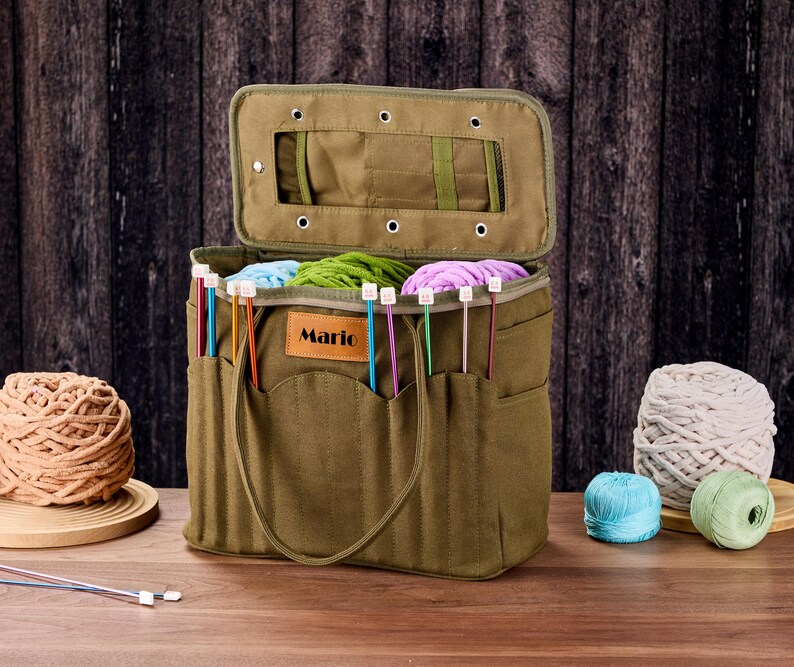 May include: An olive green canvas knitting bag with an open lid, filled with colourful yarn and knitting needles. The bag has exterior pockets and a handle. The word "Mario" is on a leather tag on the front. Yarn balls are on the table.