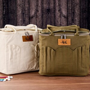 May include: Two canvas tote bags, one cream and one olive green, with top handles and front pockets. Each bag has a leather tag with a logo and a zipper closure. The olive bag has the word "Kele" on its leather tag. The bags are on a wooden surface.