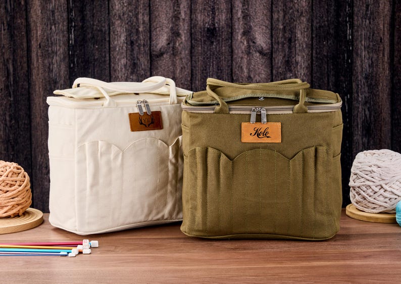 May include: Two canvas tote bags, one cream and one olive green, with top handles and front pockets. Each bag has a leather tag with a logo and a zipper closure. The olive bag has the word "Kele" on its leather tag. The bags are on a wooden surface.