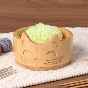 May include: A light-colored bamboo yarn bowl featuring a carved cat face design with pointed ears, closed eyes, and whiskers. The bowl contains a ball of light green yarn and is surrounded by metal crochet hooks and wooden knitting needles.