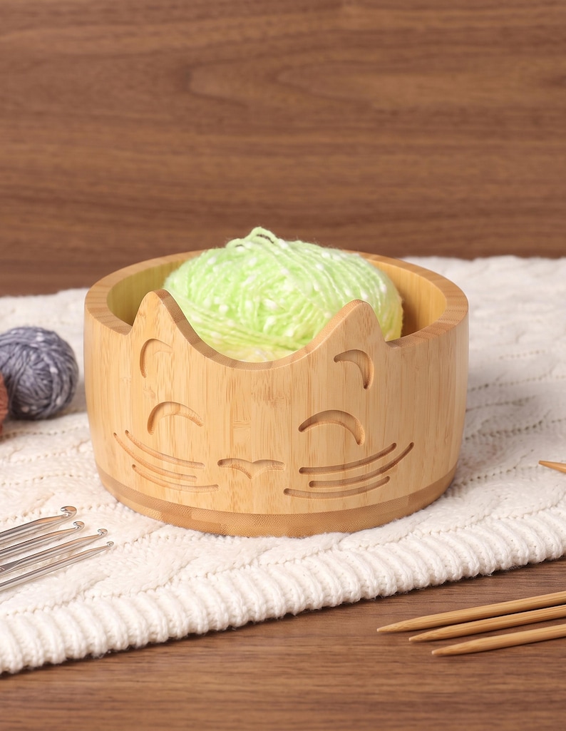 May include: A light-colored bamboo yarn bowl featuring a carved cat face design with pointed ears, closed eyes, and whiskers. The bowl contains a ball of light green yarn and is surrounded by metal crochet hooks and wooden knitting needles.