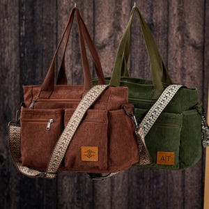Puede incluir: Dos bolsos tote de pana, uno marrón y otro verde oliva, cuelgan sobre un fondo de madera. Cada bolso tiene múltiples bolsillos delanteros, una correa para el hombro estampada y asas a juego. El bolso marrón tiene un parche de cuero con un logotipo, y el verde tiene un parche "AT".