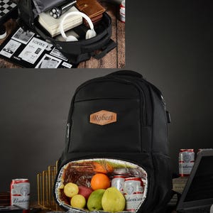 Peut inclure: Sac à dos isotherme noir avec un patch en cuir marron qui dit "Robert". Le sac à dos a un compartiment zippé et une poche en filet sur le côté. Le sac à dos est ouvert et montre une doublure isolante argentée avec des fruits et des boissons à l'intérieur.
